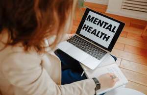 woman in white sweater taking notes with laptop that reads mental health