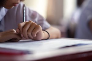 A student writes with a pencil on a piece of paper at a desk