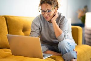 Woman with glasses researching on her laptop while sitting on a yellow coach