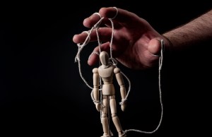 Employer manipulating the employee, emotional manipulation and obey the master concept with ominous hand pulling the strings on a marionette with moody contrast on black background